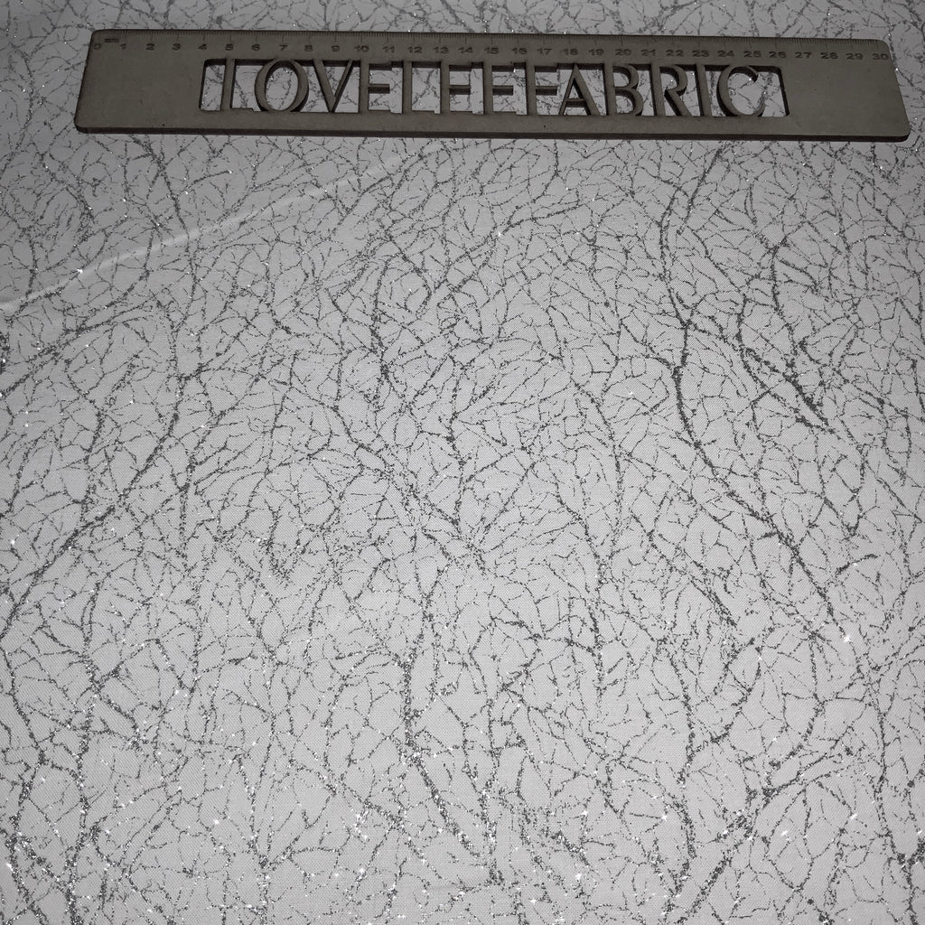 A close-up view of a white fabric with a textured silver metallic thread pattern, displaying a ruler for scale.