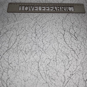 A close-up view of a white fabric with a textured silver metallic thread pattern, displaying a ruler for scale.